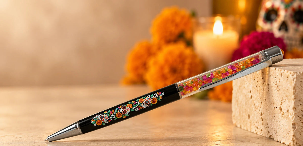 A decorative ballpoint pen with a black barrel, colorful floral patterns, and glitter-filled clear sections, resting on a stone block with a warm, festive background of marigolds and a candle.