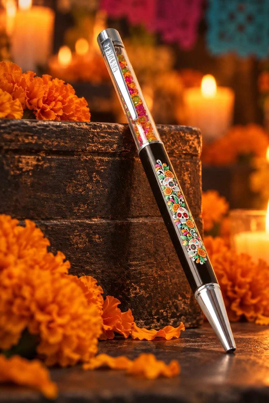 A decorative ballpoint pen with a black barrel and colorful floral patterns, resting on a wooden surface with a festive Day of the Dead background featuring papel picado banners, marigolds, and candles.