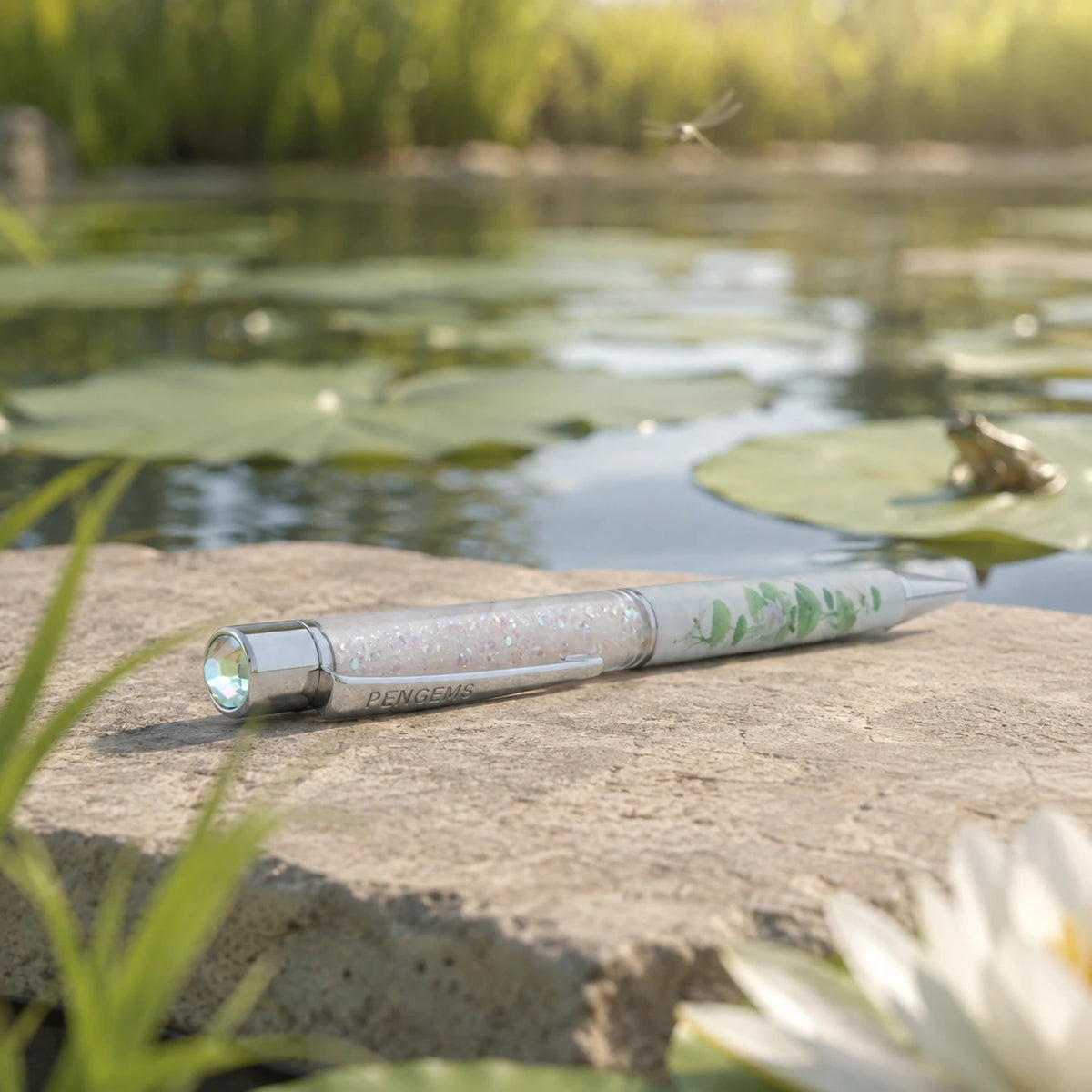 Lilypad Lounge crystal pen leaning against a rock at the edge of a pond, with a lily pad print, chrome hardware, a frog on a lily pad, and a dragonfly in flight in the background.