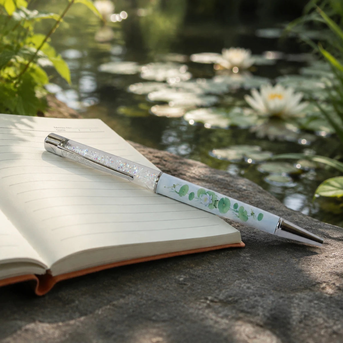 Lilypad Lounge crystal pen resting on an open notebook, with a pond and white water lilies blurred in the background.