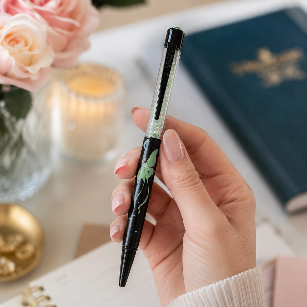 Hand holding a black pen with decorative luna moth elements on a blurred background of flowers and books.