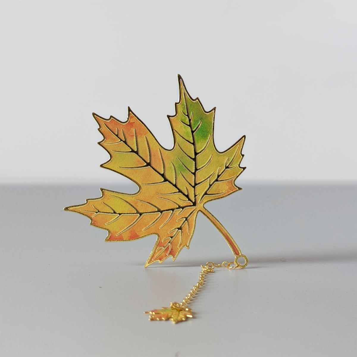 A gold-toned metal bookmark shaped like a maple leaf with autumn orange, yellow, and green enamel detailing, on a gold chain with a matching mini leaf charm.