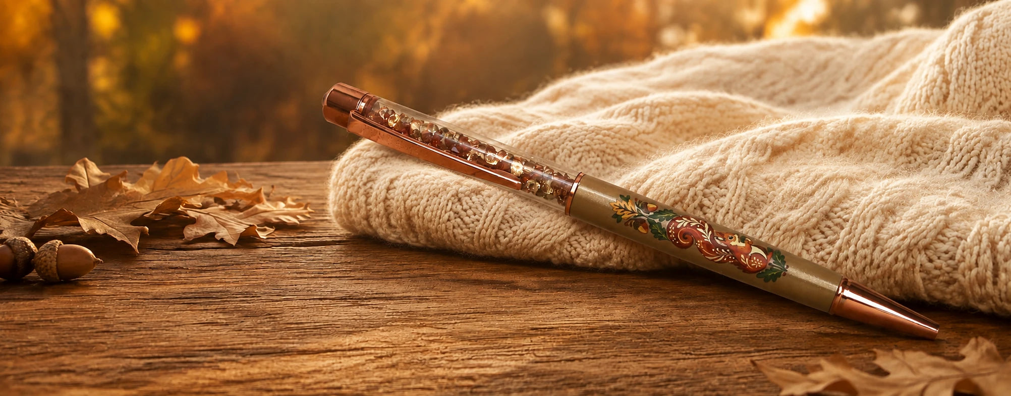 A decorative ballpoint pen with a rose gold cap and body, featuring a colorful floral illustration, resting on a beige knit blanket over a wooden table with scattered autumn leaves.