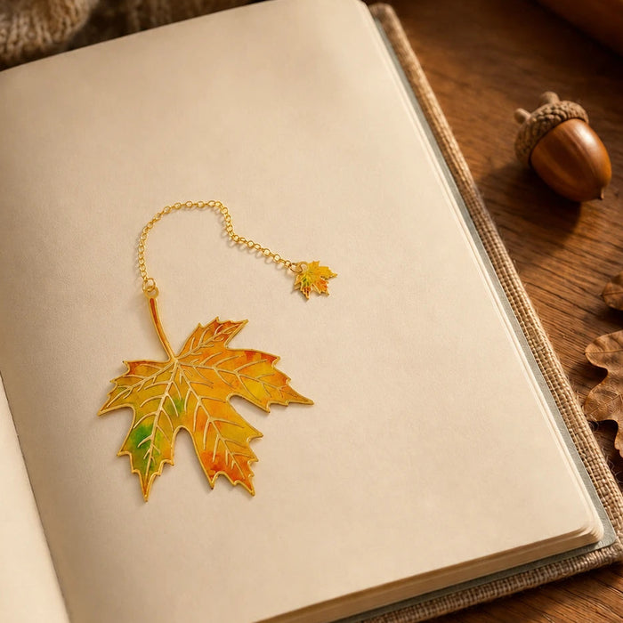 A gold-toned metal bookmark shaped like a maple leaf with autumn yellow, orange, and green enamel detailing, resting on the inside page of an open book. A knitted beige scarf, a wooden acorn, and dried leaves are visible around it on a wooden surface.