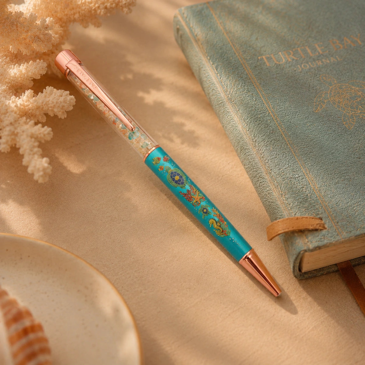 PenGems - Turtle Bay - crystal pen beside a teal journal embossed with a gold sea turtle, coral branch nearby, rose gold hardware catching the light.